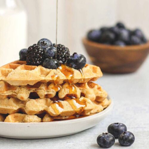 Syrup being poured over a stack of waffles topped with fresh blackberries and blueberries.