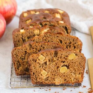 Slices of healthy apple bread on a rectangular wire cooling rack with a gold knife on the side and apples in the background.