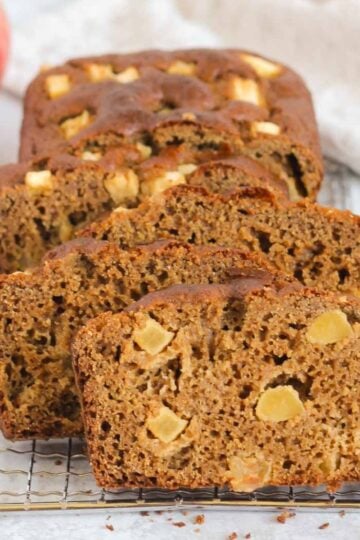 Slices of healthy apple bread on a rectangular wire cooling rack with a gold knife on the side and apples in the background.
