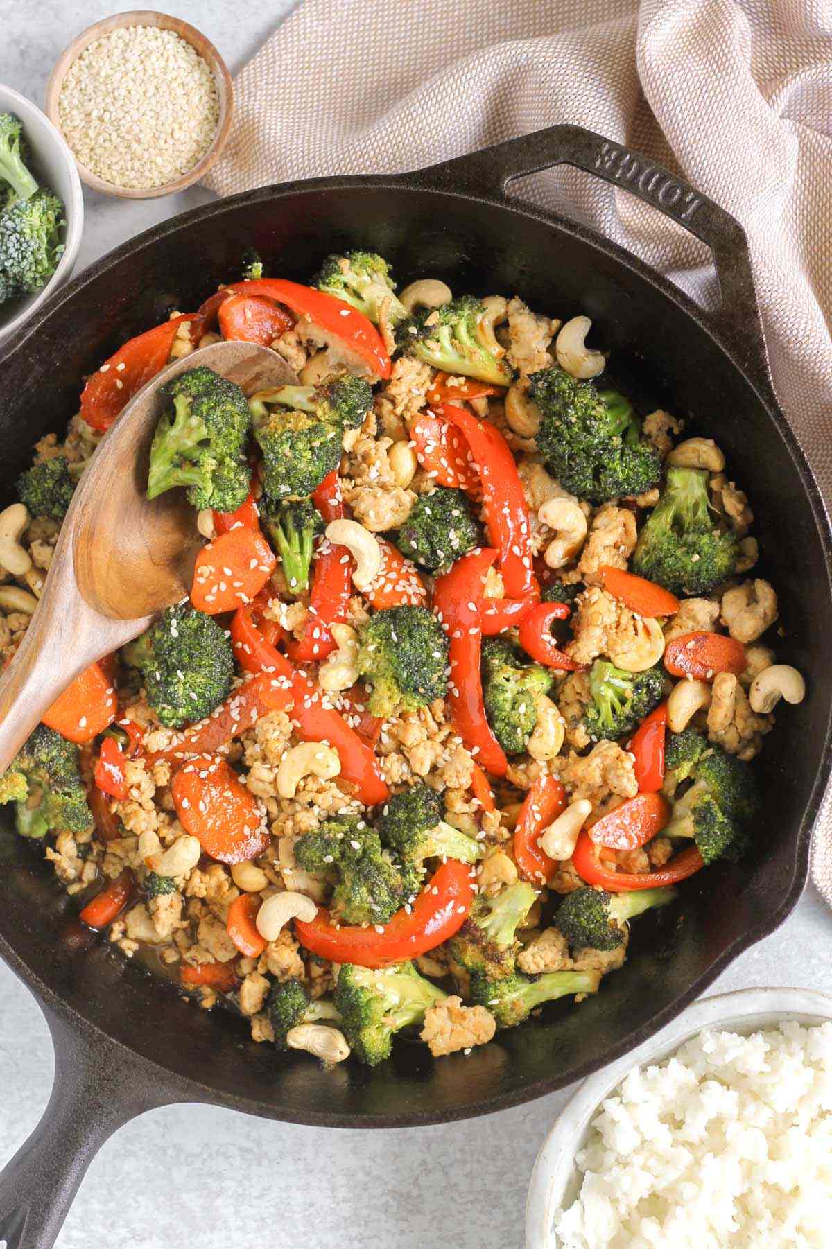 Ground chicken stir fry sprinkled with sesame seeds and cashews in a cast iron skillet with a wooden serving spoon scooping some out.