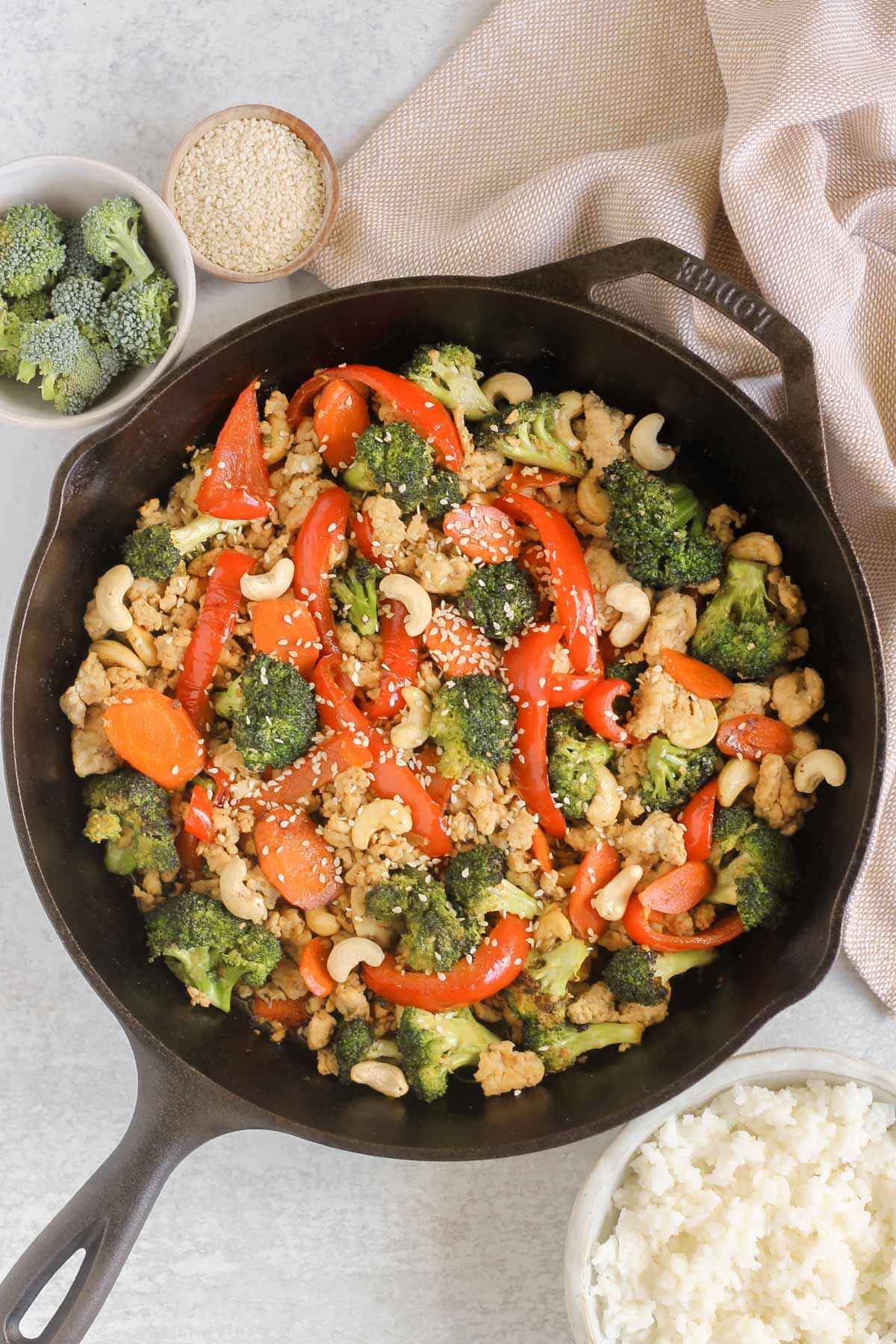 Ground chicken and veggie stir fry in a cast iron skillet with a bowl of broccoli and cooked rice on the side.