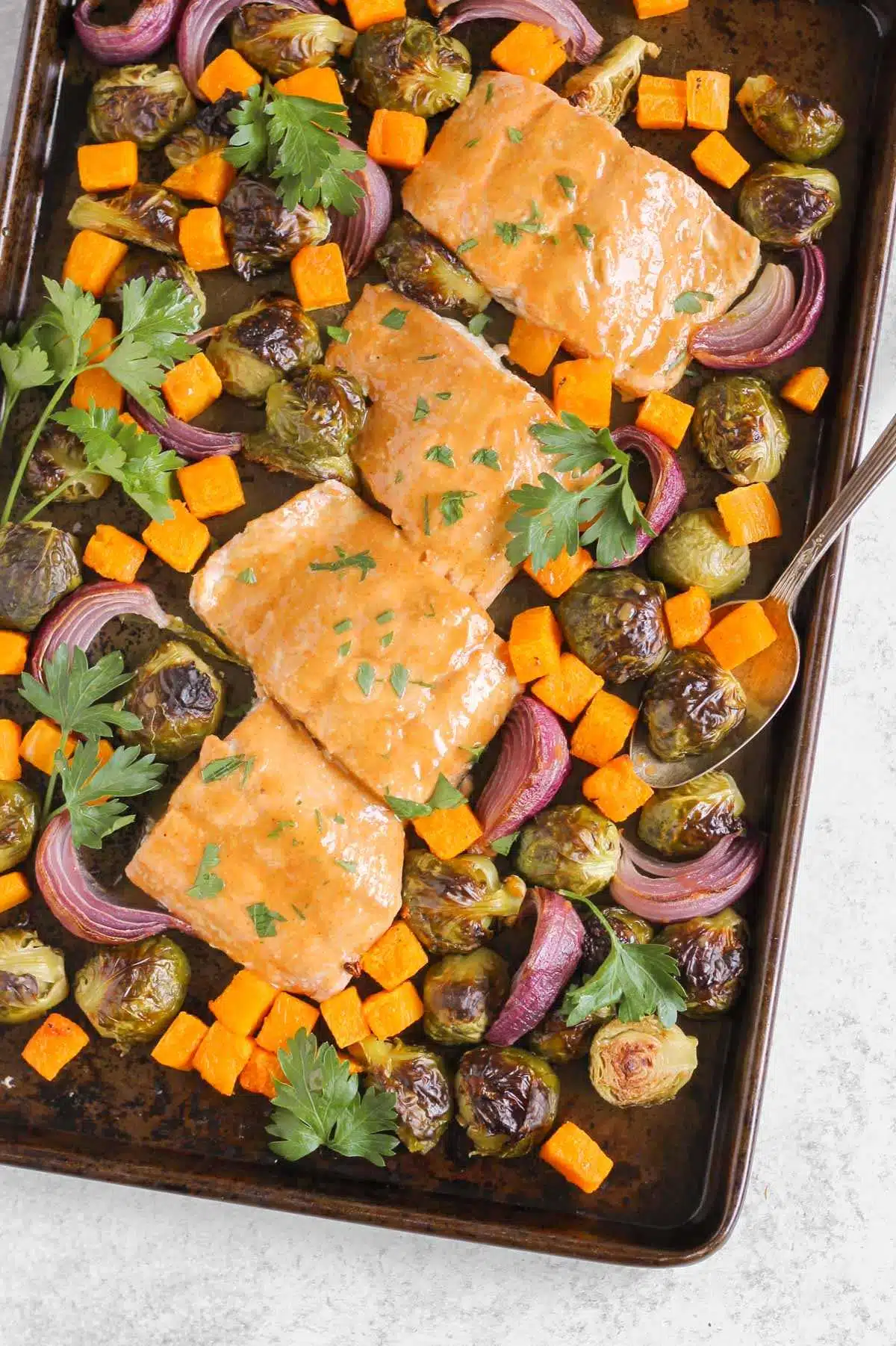 Roasted salmon and veggies on a baking sheet with parsley sprinkled around and a vintage spoon scooping up some of the veggies.