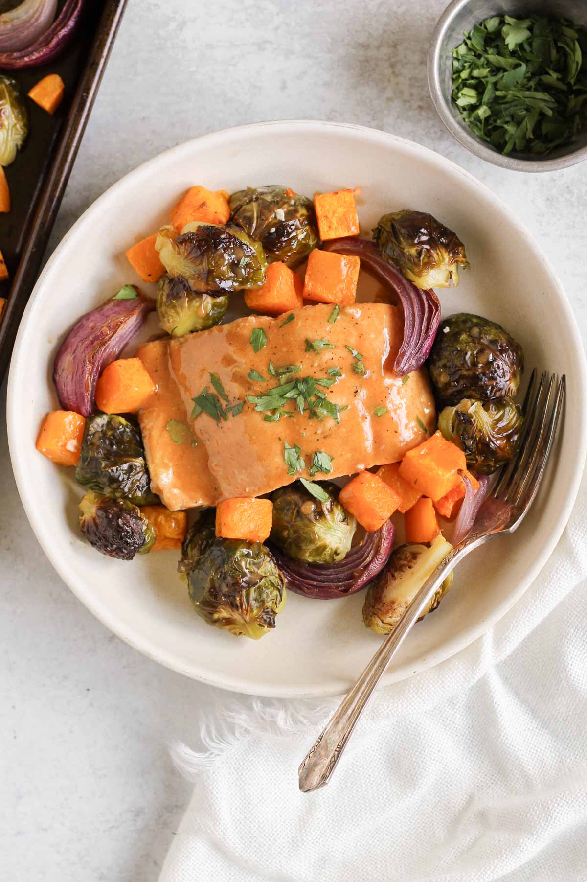 Single salmon fillet sprinkled with parsley with roasted veggies around the filet sitting in a shallow single serve bowl.