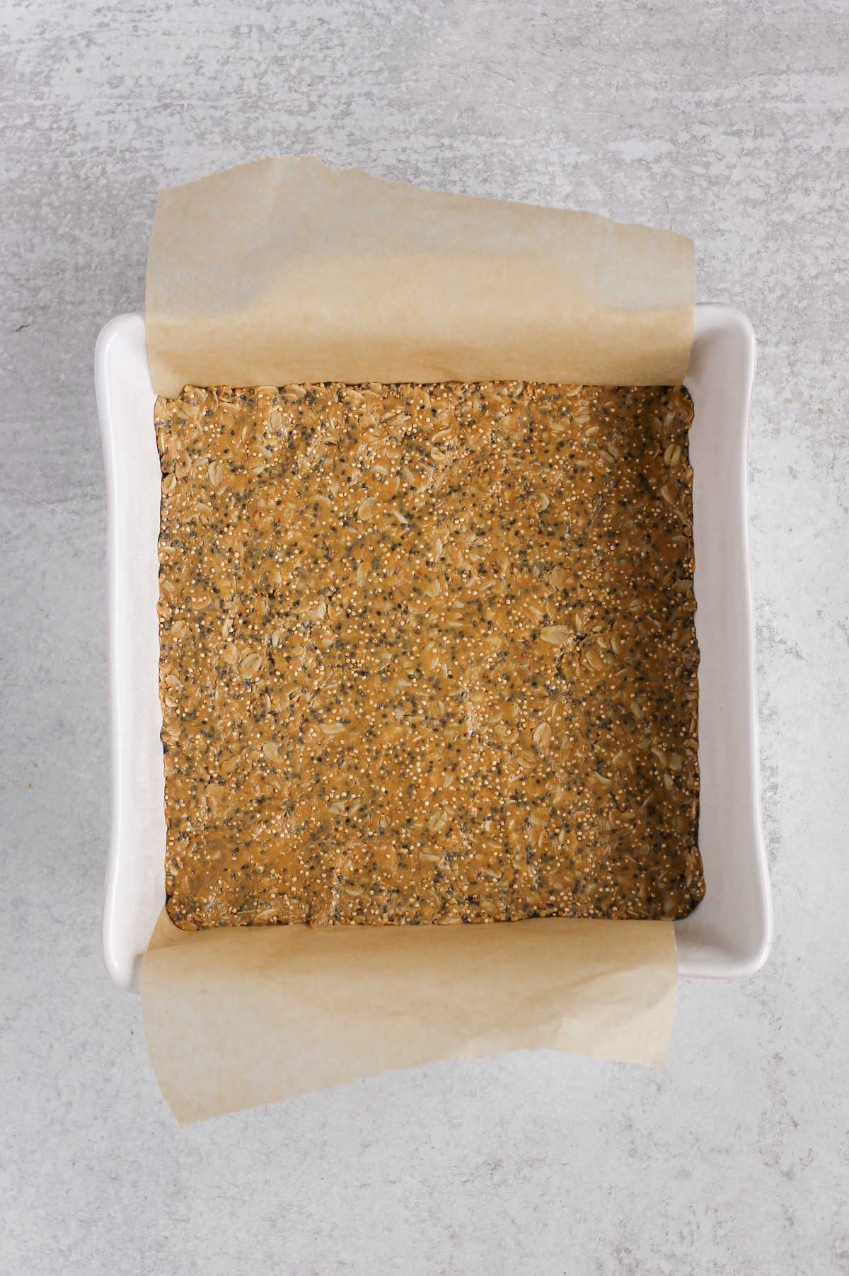 Protein granola bar mix pressed into the bottom of an 8x8 baking dish with parchment paper hanging over the sides.