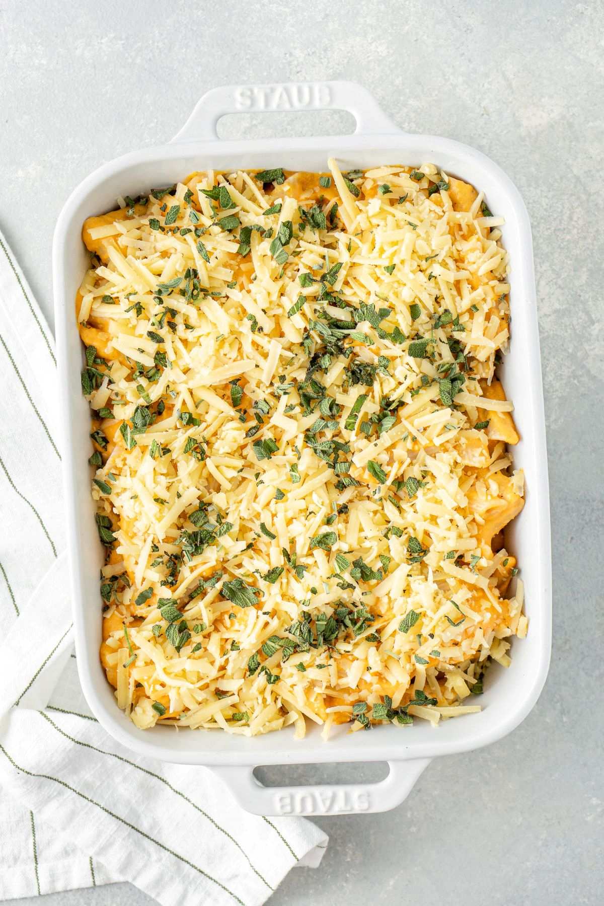 Butternut squash mac and cheese topped shredded cheese and fresh sage in a rectangular baking dish before being baked.