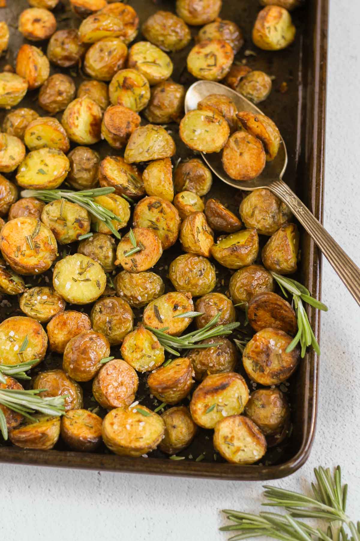 Roasted sliced baby potatoes on a baking sheet with sprigs of fresh rosemary and a vintage serving spoon.