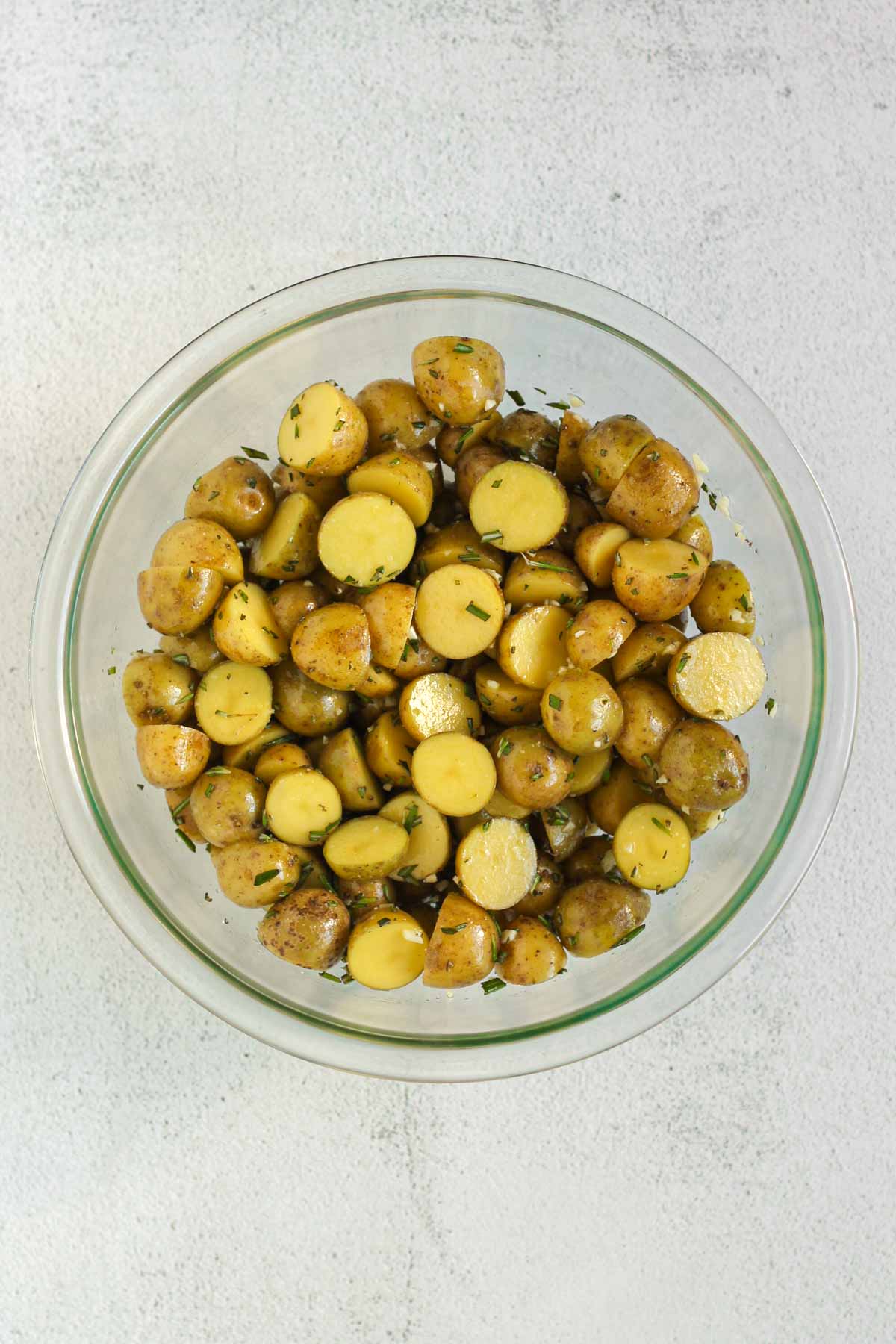 Sliced baby potatoes tossed with olive oil, rosemary and garlic in a small glass mixing bowl.
