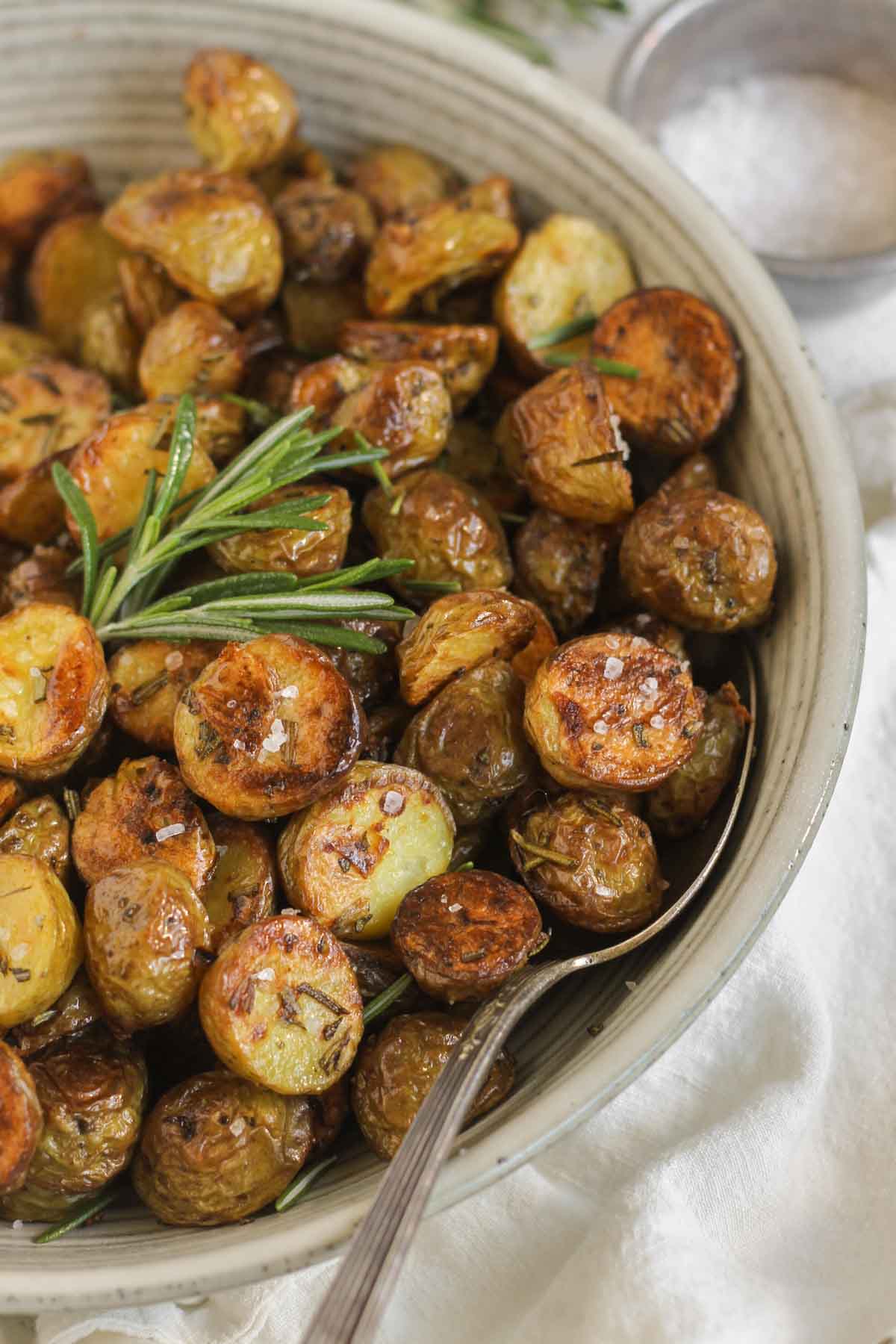 Crispy roasted potatoes sprinkled with sea salt with a sprig of fresh rosemary and a serving spoon in a small serving bowl.