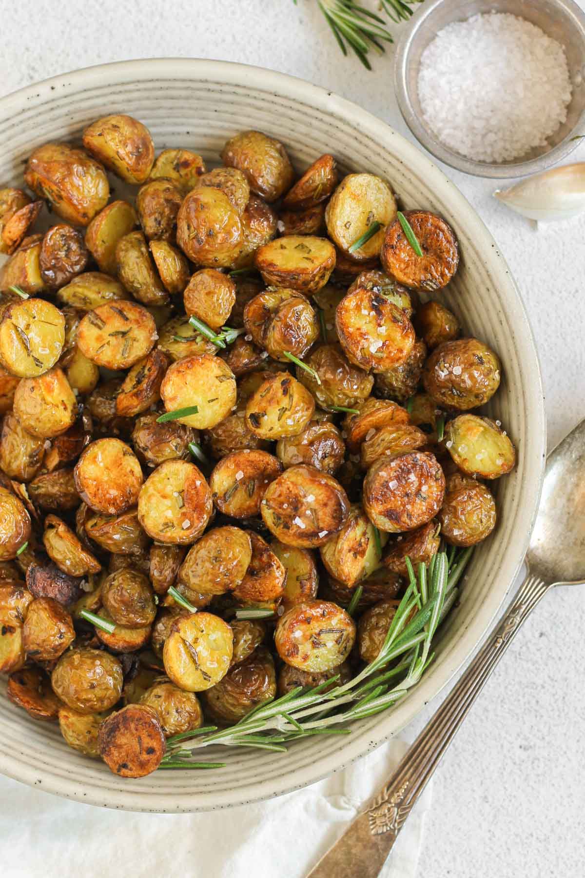 Serving bowl with roasted baby potatoes with a sprig of fresh rosemary and small ramekin of salt off to the side.