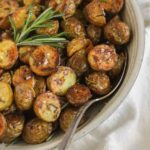 Crispy roasted potatoes sprinkled with sea salt with a sprig of fresh rosemary and a serving spoon in a small serving bowl.