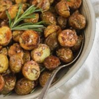 Crispy roasted potatoes sprinkled with sea salt with a sprig of fresh rosemary and a serving spoon in a small serving bowl.