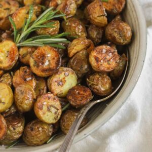 Crispy roasted potatoes sprinkled with sea salt with a sprig of fresh rosemary and a serving spoon in a small serving bowl.