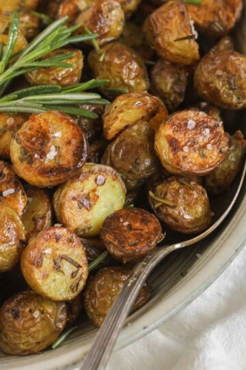 Crispy roasted potatoes sprinkled with sea salt with a sprig of fresh rosemary and a serving spoon in a small serving bowl.