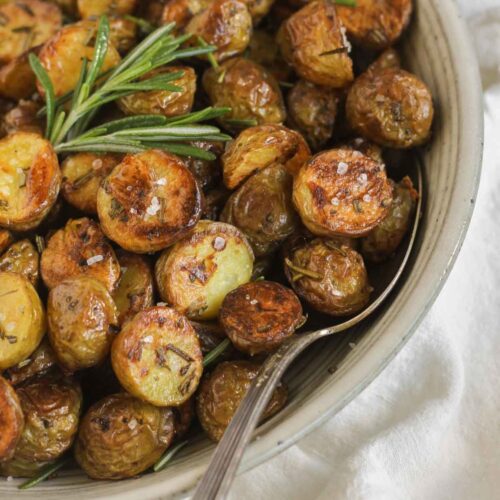 Crispy roasted potatoes sprinkled with sea salt with a sprig of fresh rosemary and a serving spoon in a small serving bowl.
