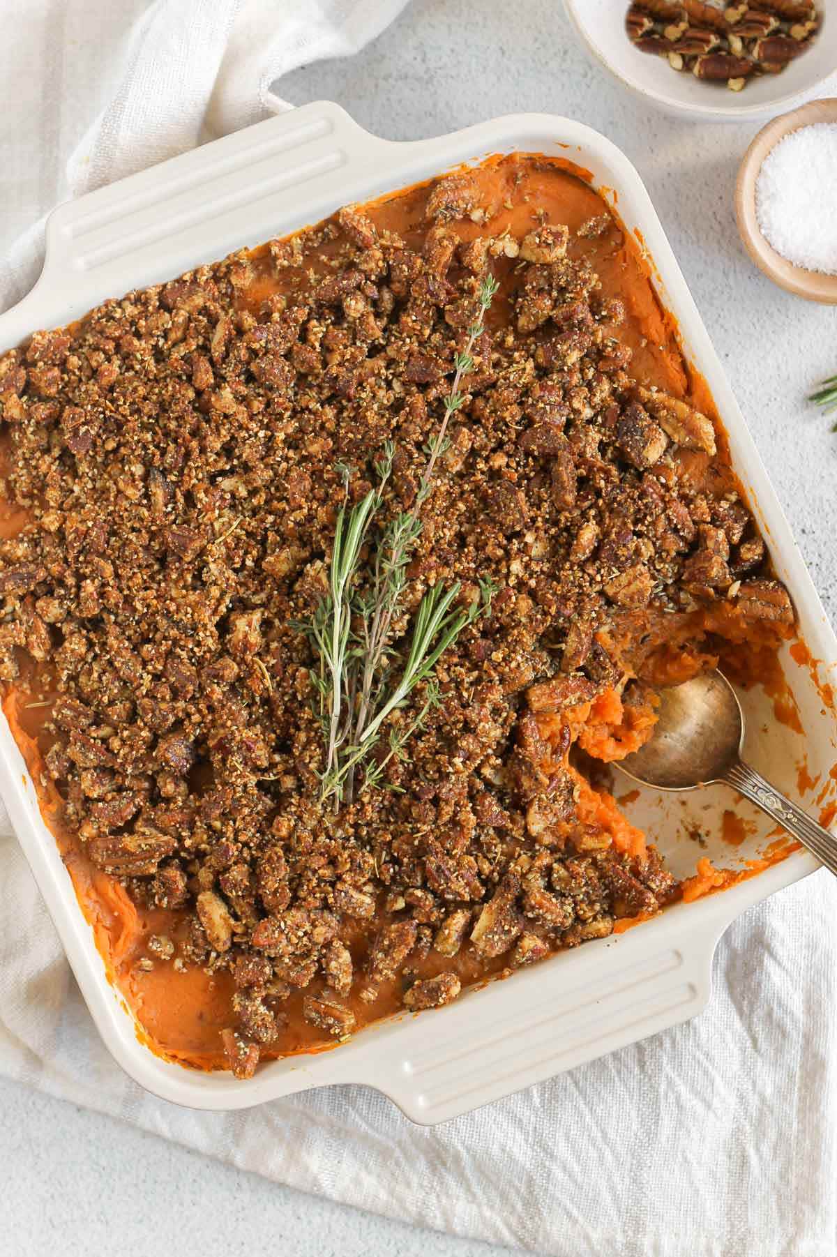 Sweet potato baked covered with pecan crumble and fresh herbs in a square baking dish with a serving spoon scooping some out of the corner.