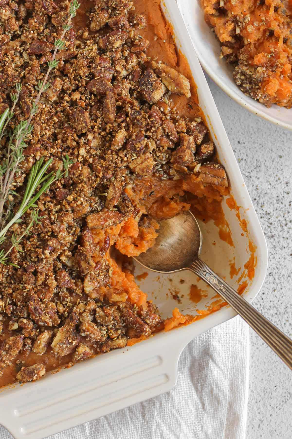 Sweet potato baked covered with pecan crumble and fresh herbs in a square baking dish with the corner serving scooped out and a serving spoon in the dish.