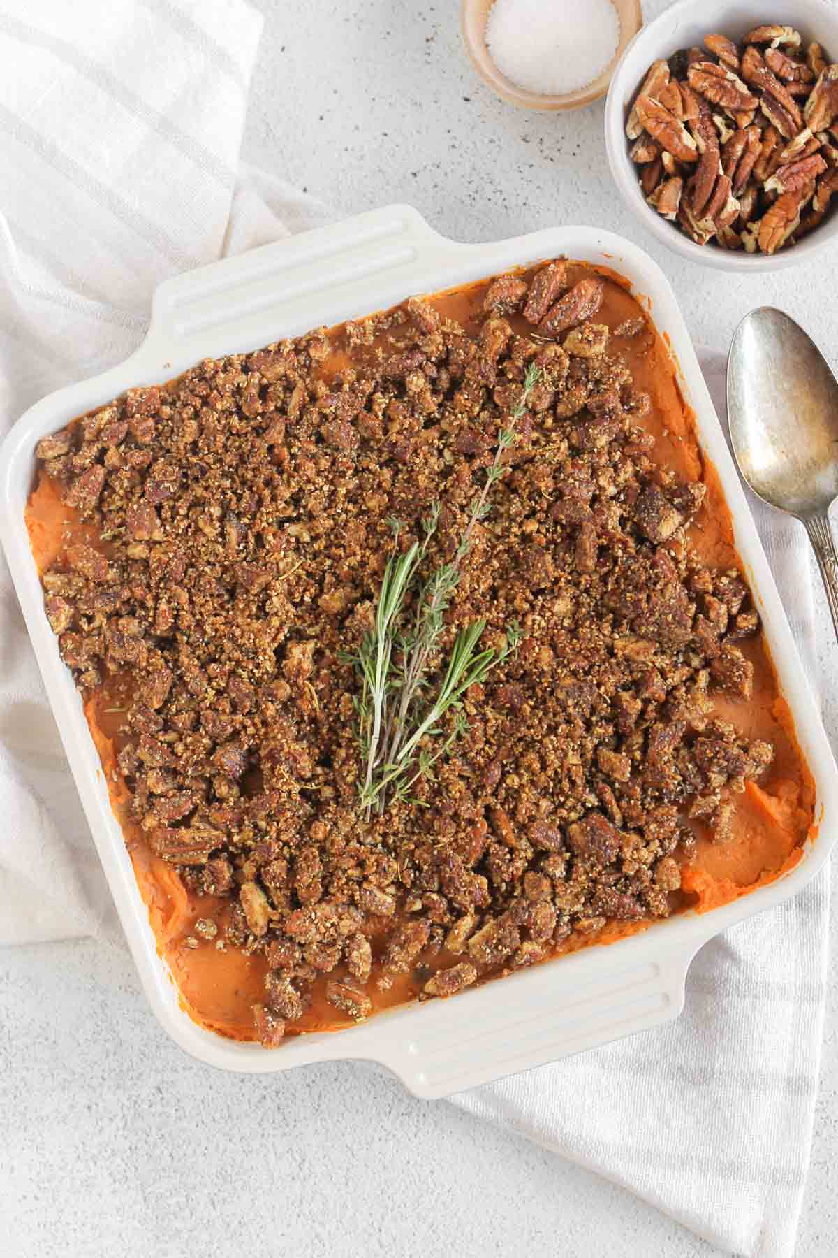 Sweet potato baked covered with pecan crumble and fresh herbs in a square baking dish with a serving spoon and a bowl of pecans on the side.