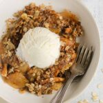 Single serving of apple crisp in a small bowl topped with a scoop of vanilla ice cream with 2 vintage forks in the bowl.