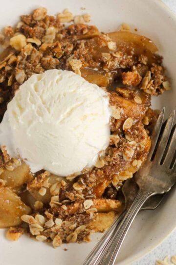 Single serving of apple crisp in a small bowl topped with a scoop of vanilla ice cream with 2 vintage forks in the bowl.