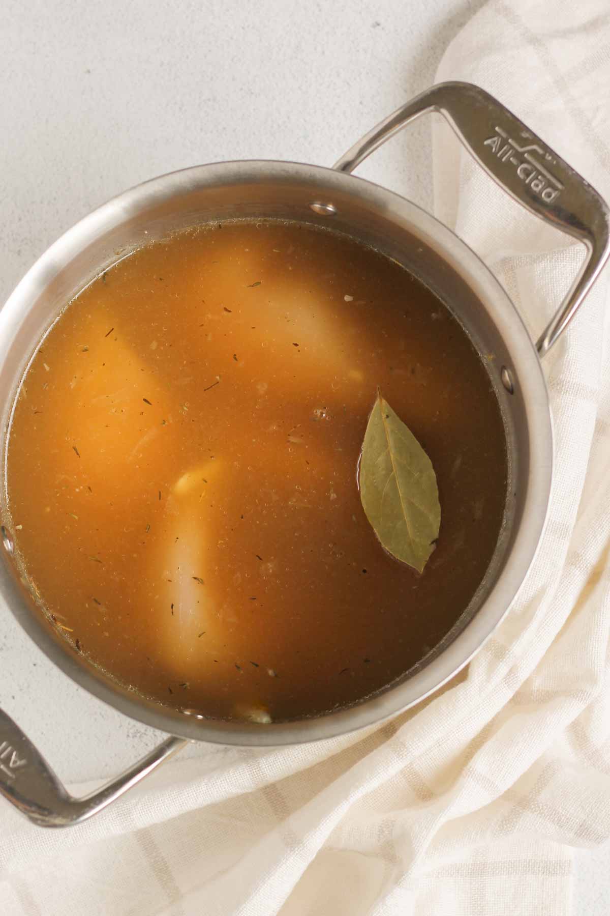 Chicken breasts covered with broth and a bay leaf in a stainless steel stock pot.