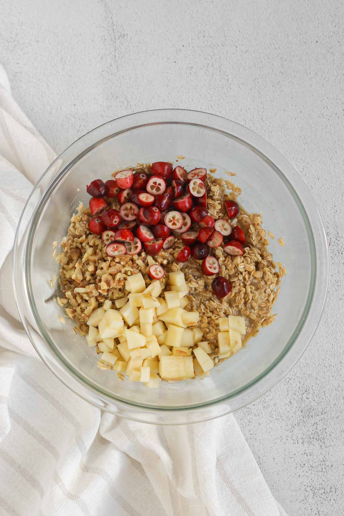 Oatmeal breakfast cookie dough in a glass mixing bowl with cranberries, apples, and walnuts added to the top.