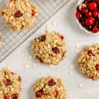 Multiple cranberry apple breakfast cookies spread out over a surface and cooling rack with a small dish of cranberries on the side.