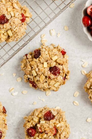 Multiple cranberry apple breakfast cookies spread out over a surface and cooling rack with a small dish of cranberries on the side.