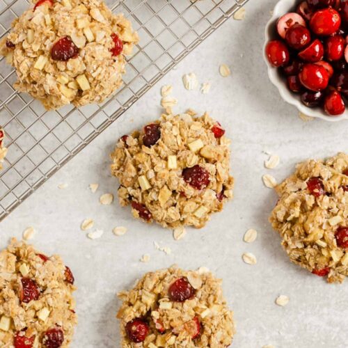 Multiple cranberry apple breakfast cookies spread out over a surface and cooling rack with a small dish of cranberries on the side.