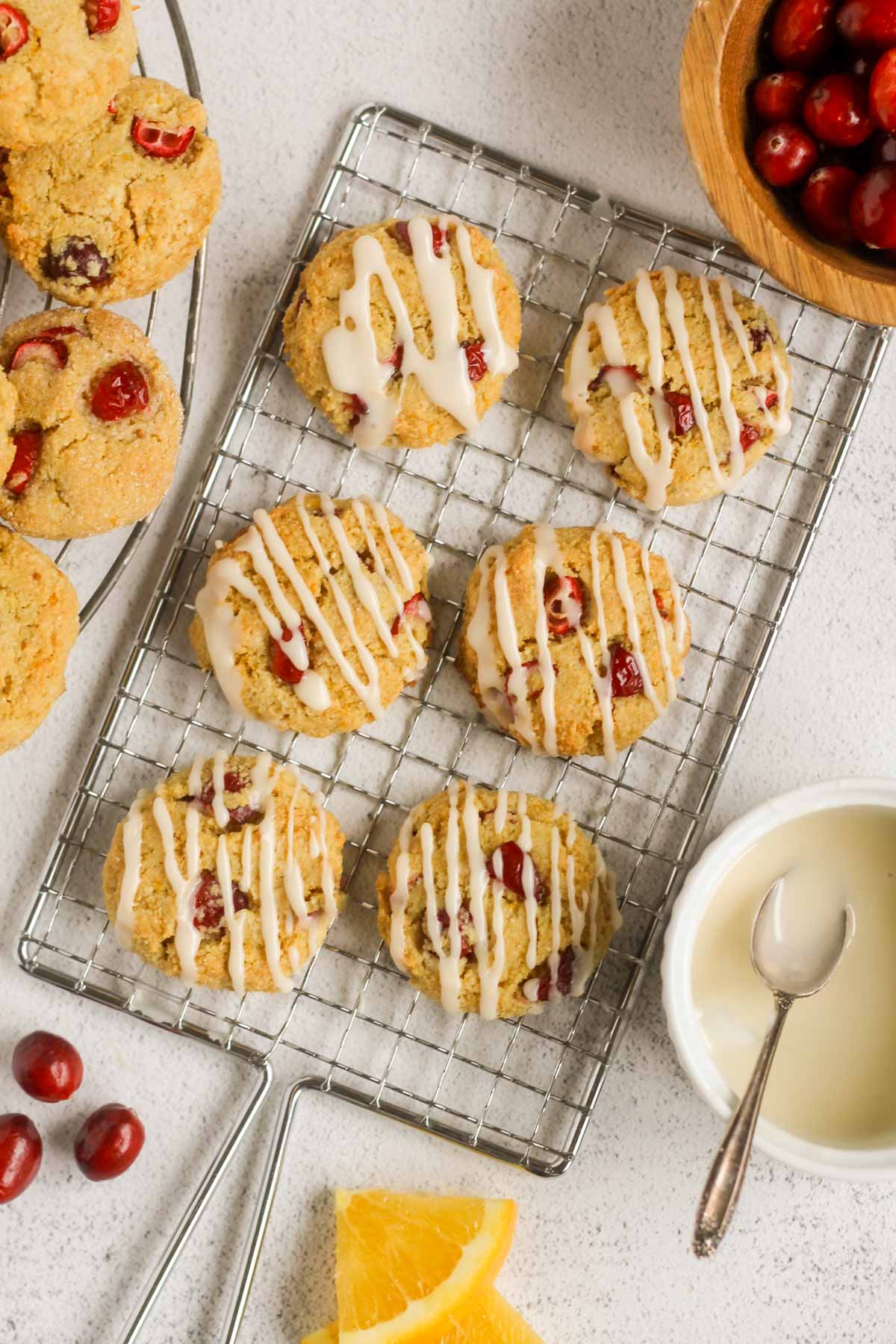Cranberry almond flour cookies drizzled with icing on rectangular cooking rack with a small bowl of icing and a bowl of cranberries on the side.