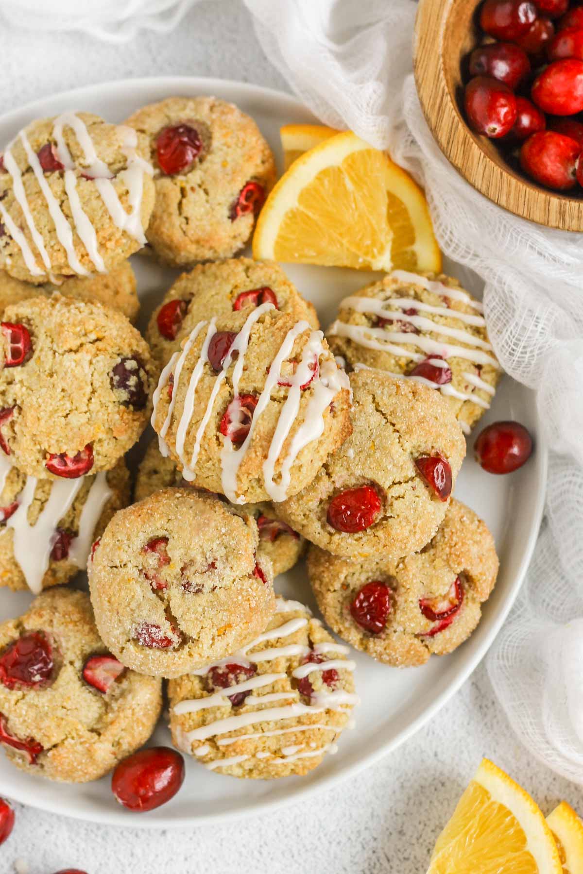 Cranberry almond flour cookies on a plate, so drizzled in icing and some topped with sugar, with a bowl of cranberries and orange slices on the side.