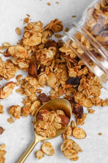 Oats and honey granola spilled from a mason jar with a gold spoon in the spilled granola.