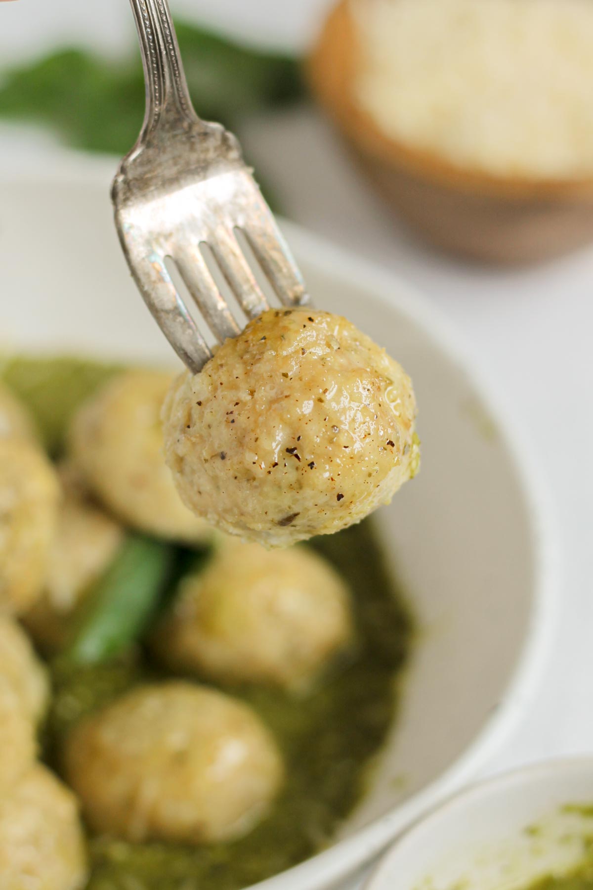 A single gluten-free chicken meatball in a fork with a bowl of meatballs in the background.