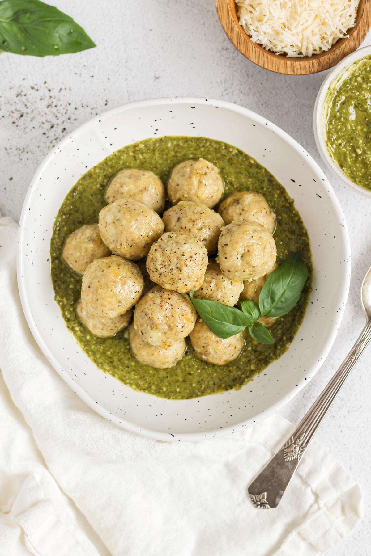 Chicken meatballs stacked in a bowl with pesto with small bowls of pesto and shredded parmesan cheese on the side.