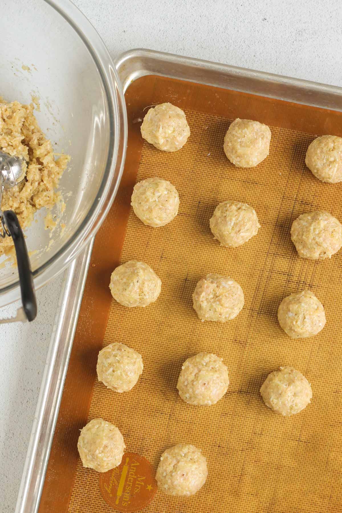 Raw ground chicken meatballs on a baking sheet with a bowl of ground chicken mix on the side.