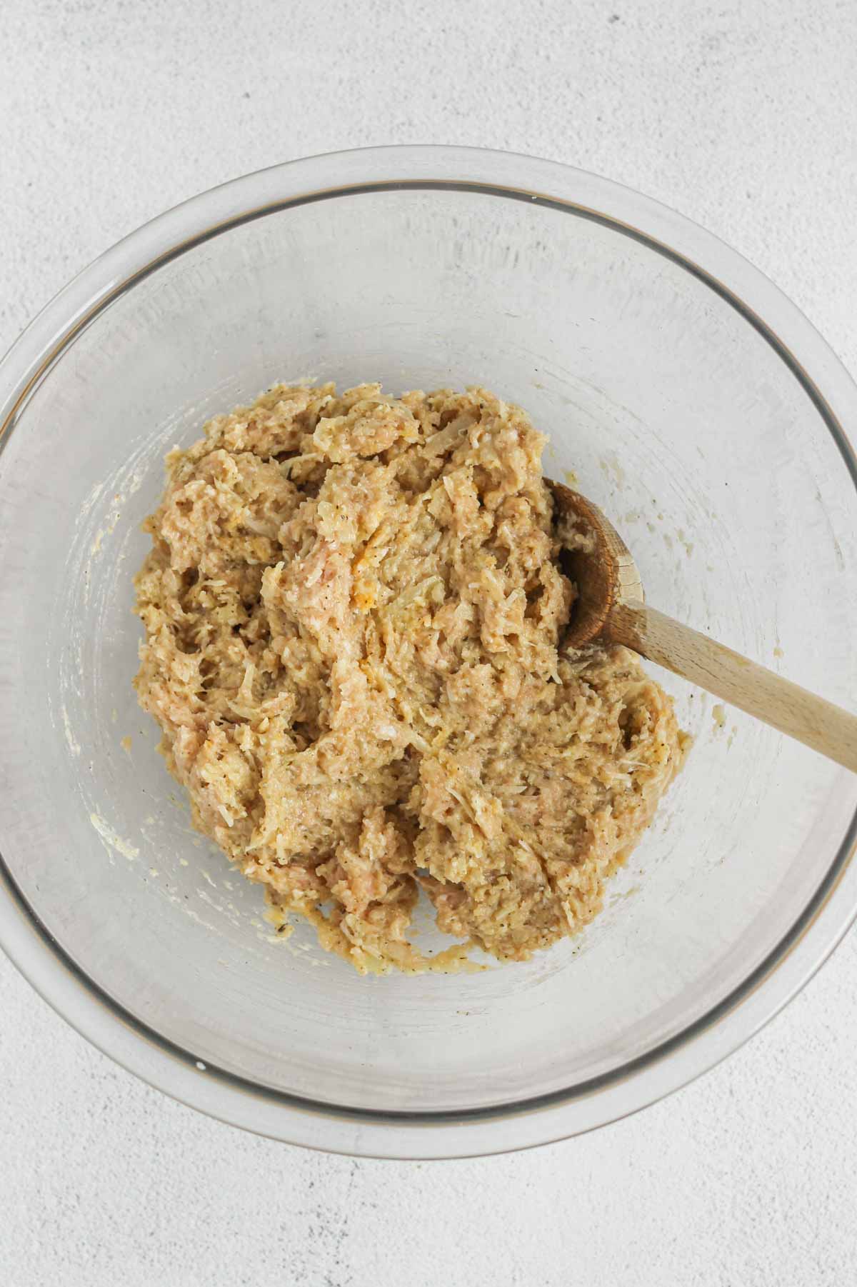 Ground chicken mixed with an egg and seasoning in a glass mixing bowl with a wooden spoon.