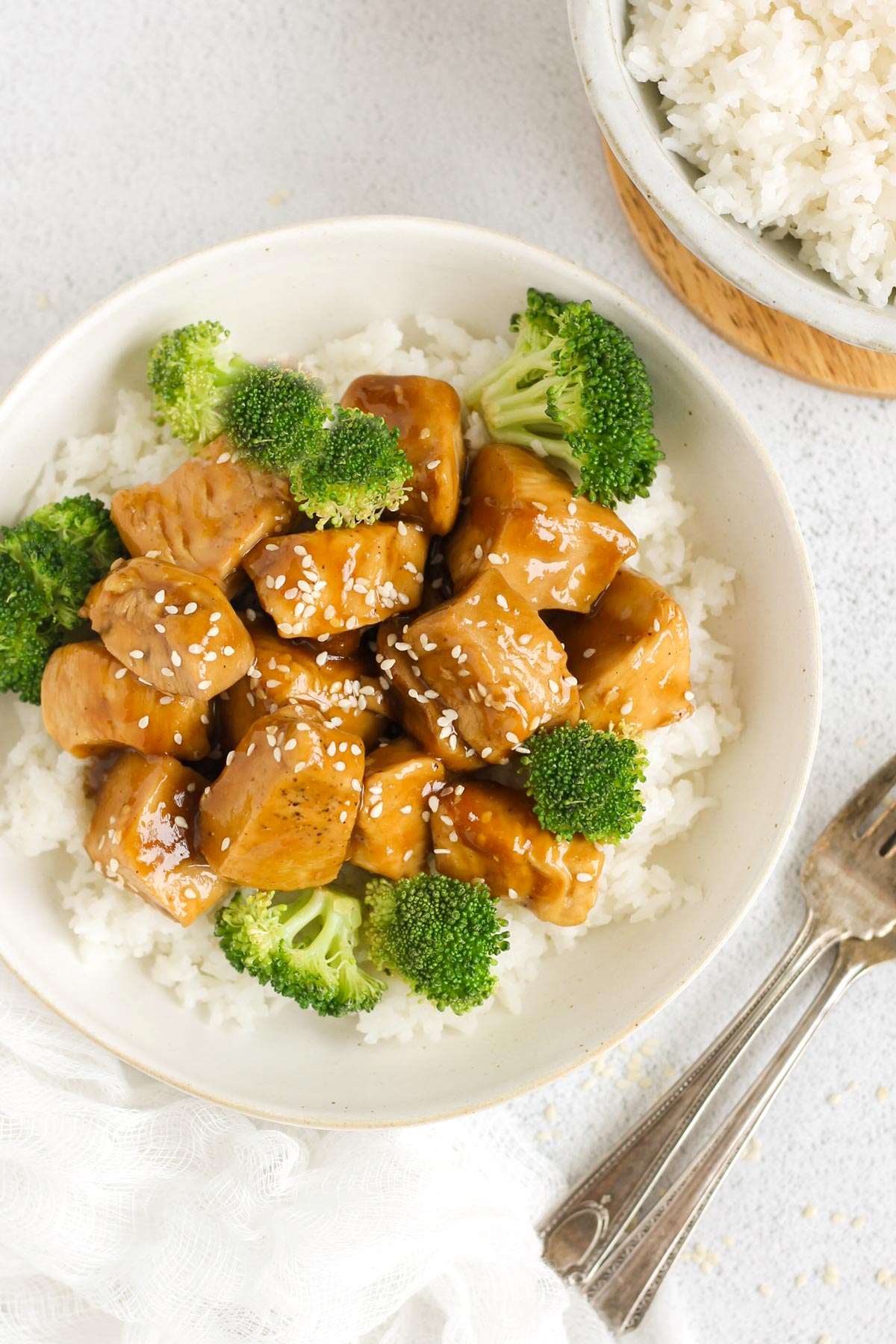 Teriyaki chicken pieces with broccoli on a bed of rice in a single serve shallow bowl with 2 forks on the side and a bowl of rice in the background.
