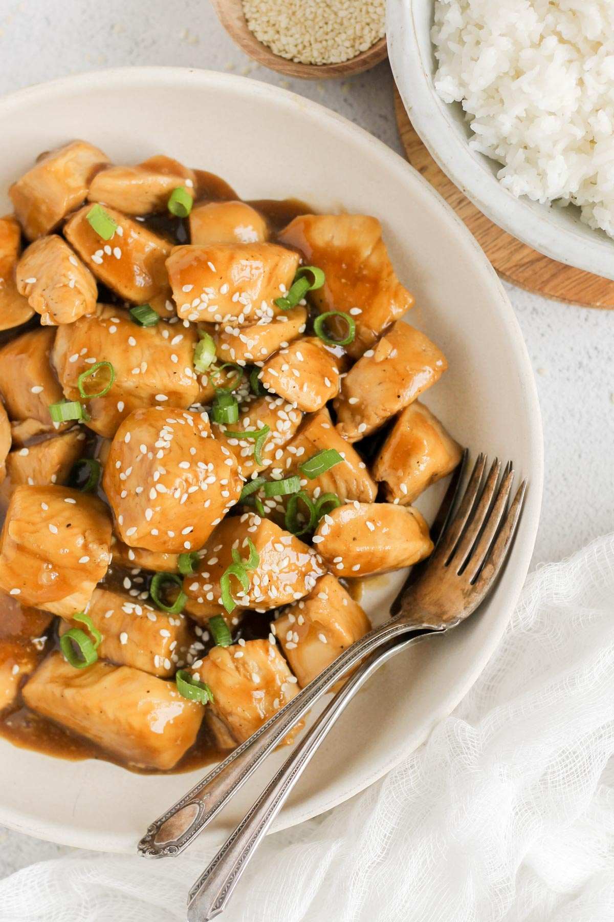 Teriyaki chicken pieces topped with sliced green onions and sesame seeds in a serving bowl with two forks in the bowl and a bowl of rice on the side.