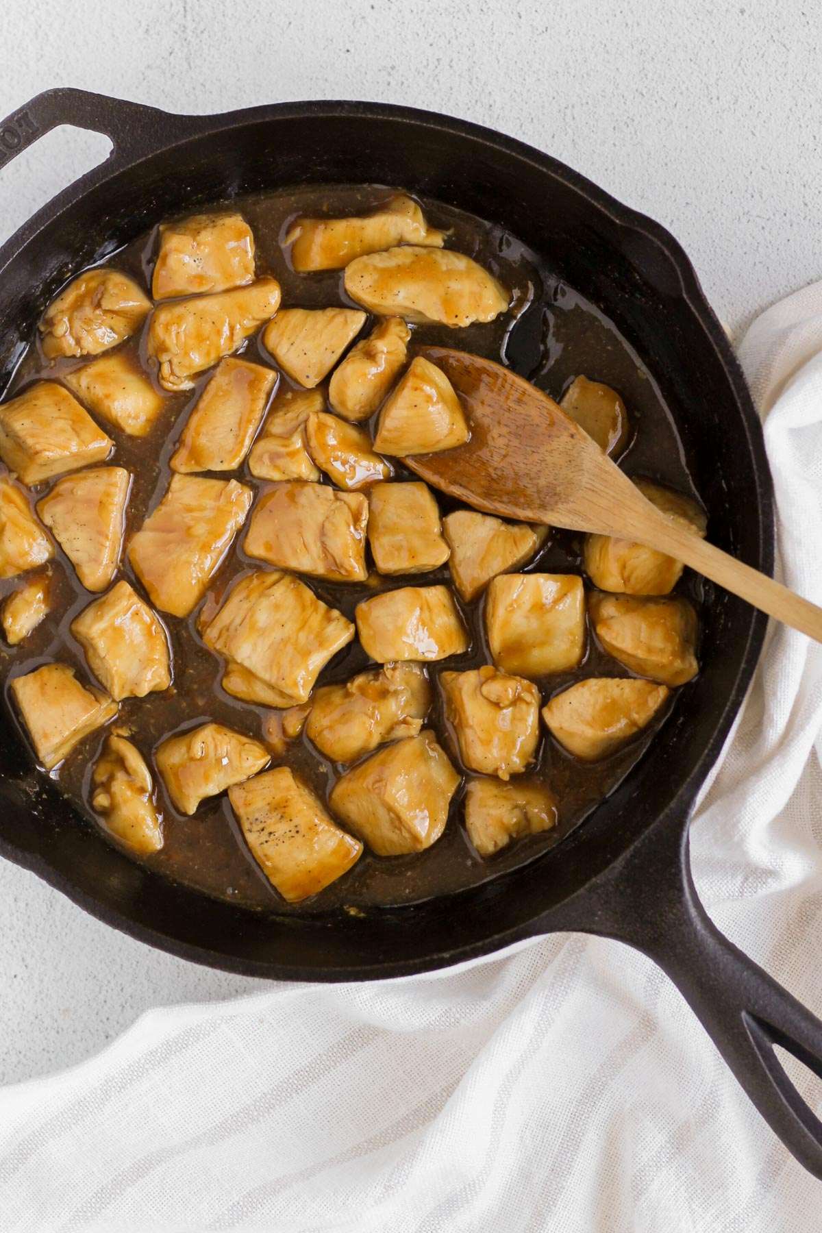 Chicken pieces with teriyaki sauce in a cast iron skillet with a wooden spoon scooping up a piece of chicken.