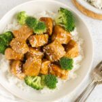 Teriyaki chicken pieces with broccoli on a bed of rice in a single serve shallow bowl with 2 forks on the side.