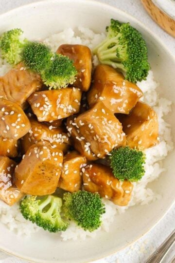 Teriyaki chicken pieces with broccoli on a bed of rice in a single serve shallow bowl with 2 forks on the side.
