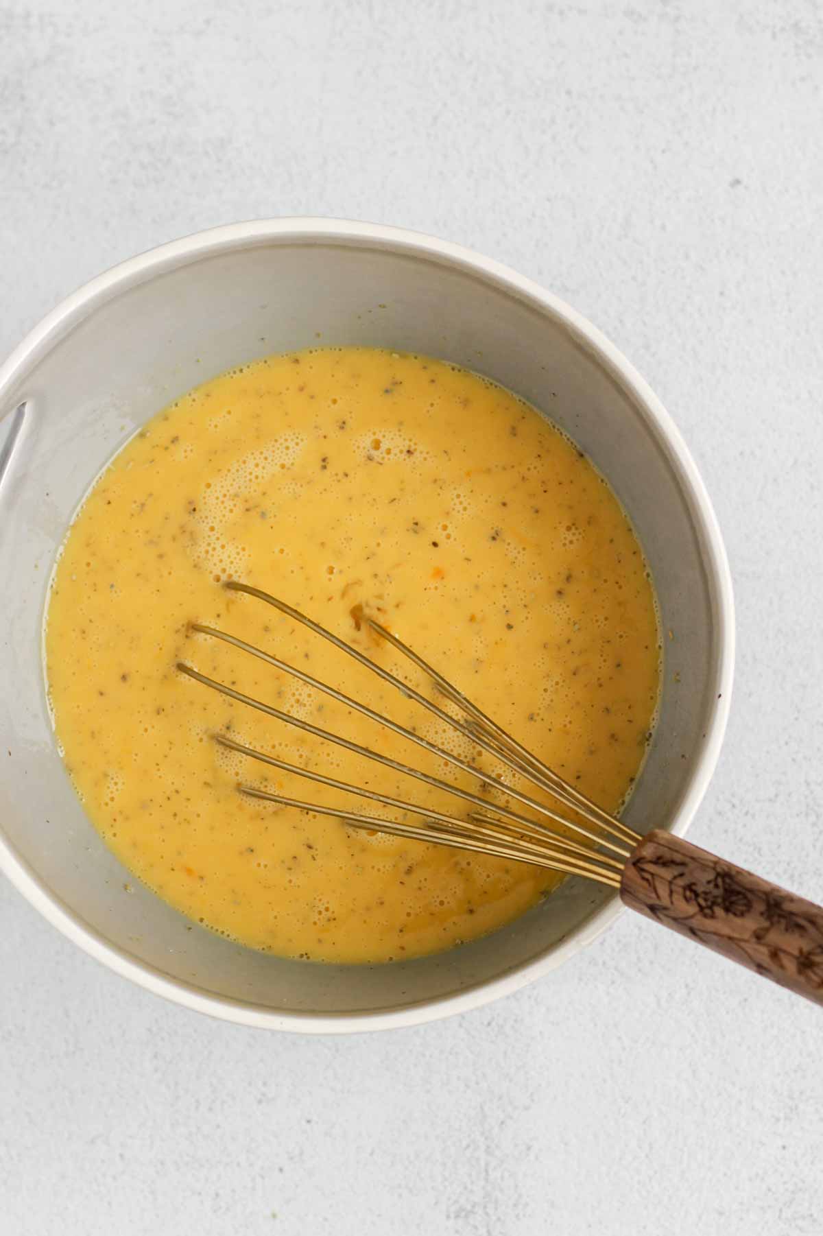 Eggs and seasoning whisked in a mixing bowl with a whisk in the bowl.