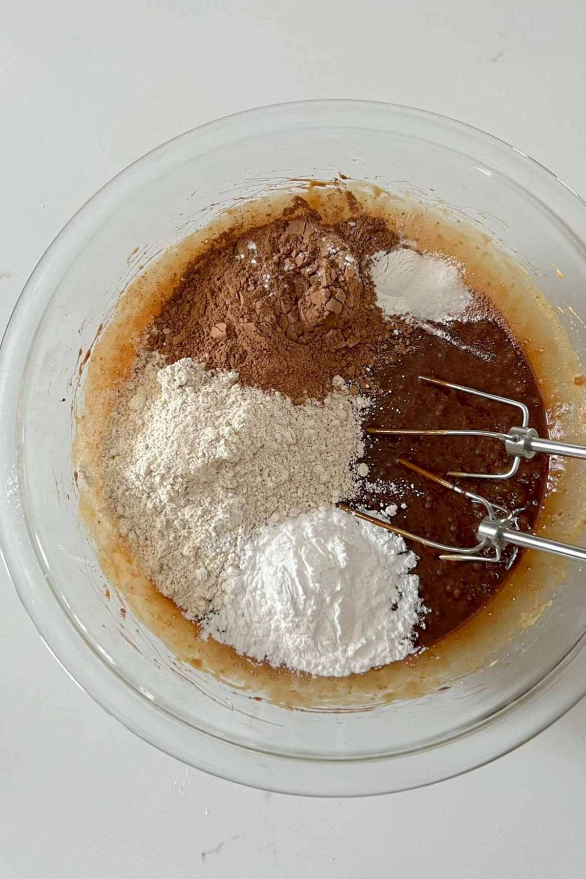 Brownie batter wet ingredients in a glass mixing bowl with separate piles of dry ingredients added to the bowl.