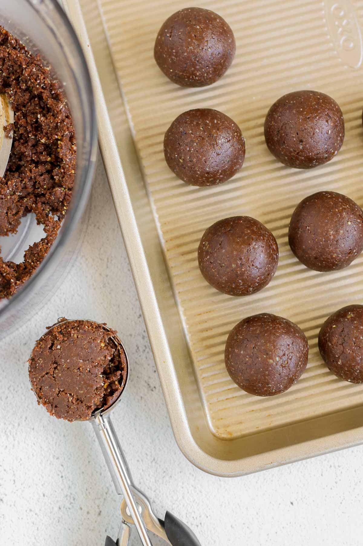 Brownie balls on a cookie sheet with a spring-loaded scoop filled with brownie ball dough next to the cookie sheet.