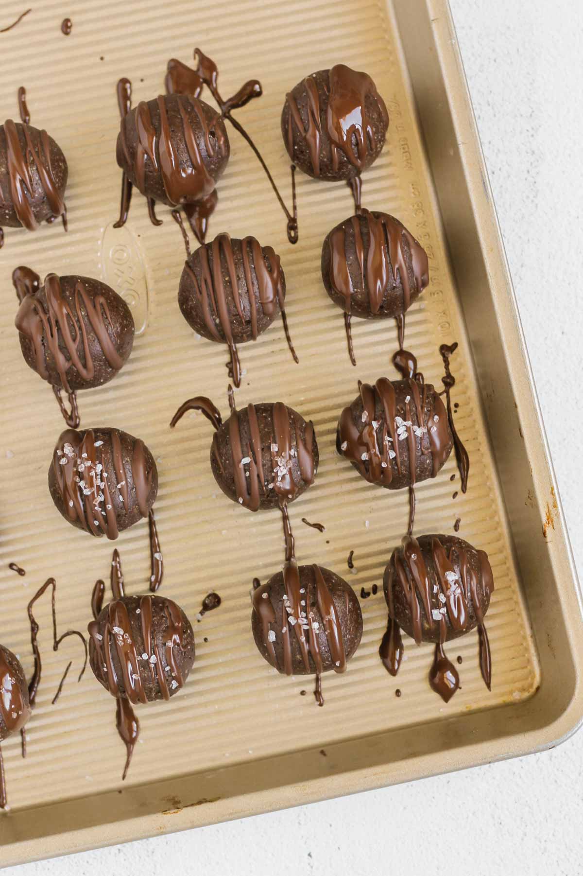 Protein brownie balls drizzled with chocolate and some sprinkled with sea salt on a cookie sheet.