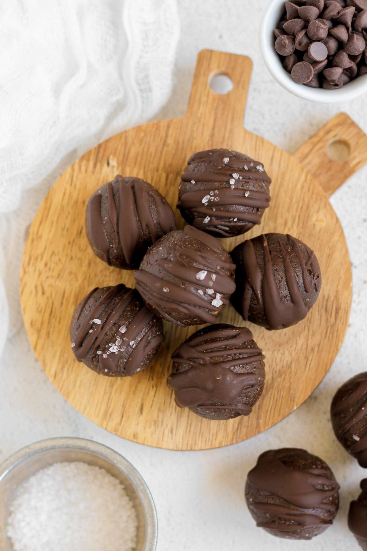 Six chocolate brownie balls on a small wood tray with additional balls and bowl of chocolate chips on the side.