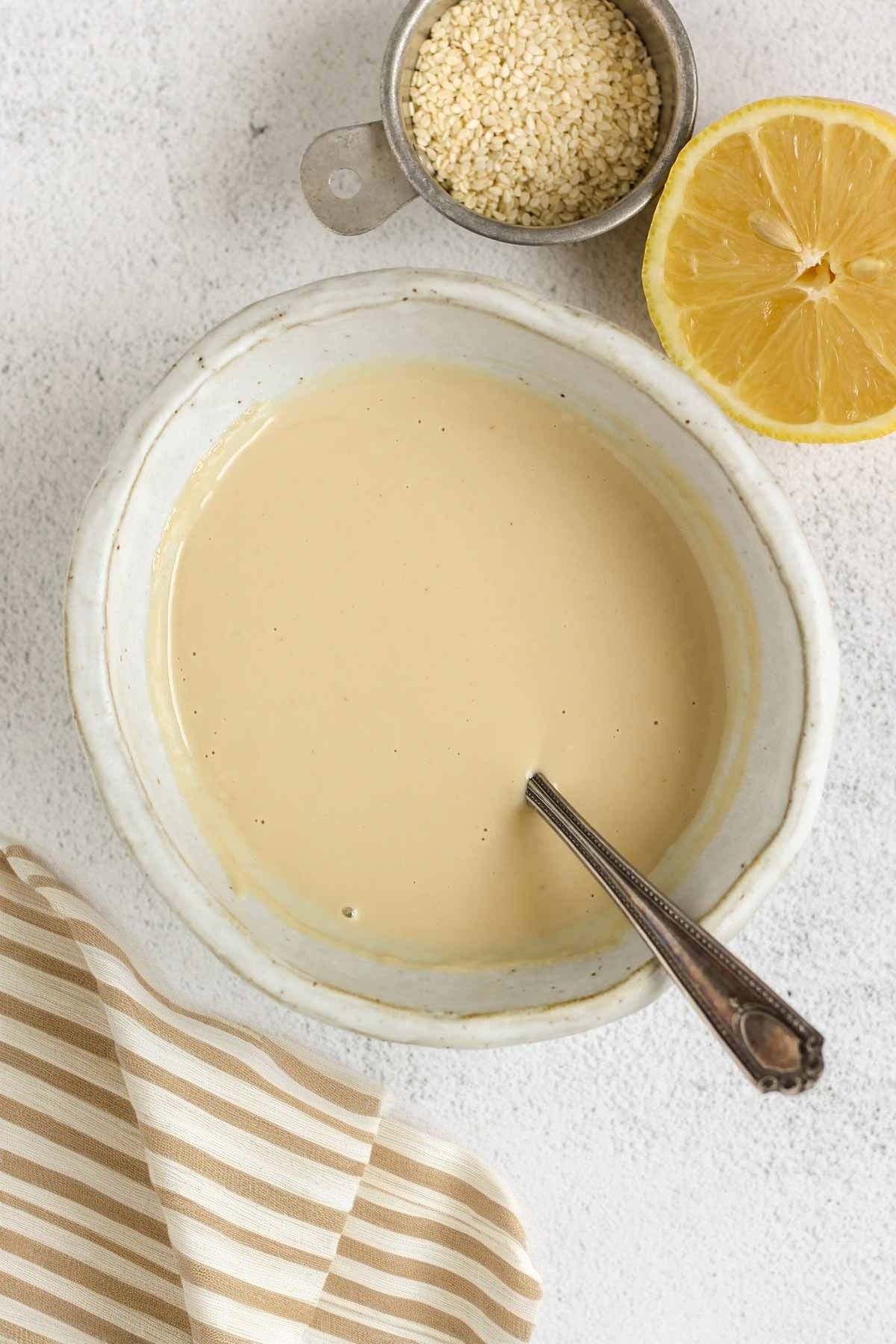Maple tahini dressing in a shallow bowl with a dish of sesame seeds and half a lemon on the side.