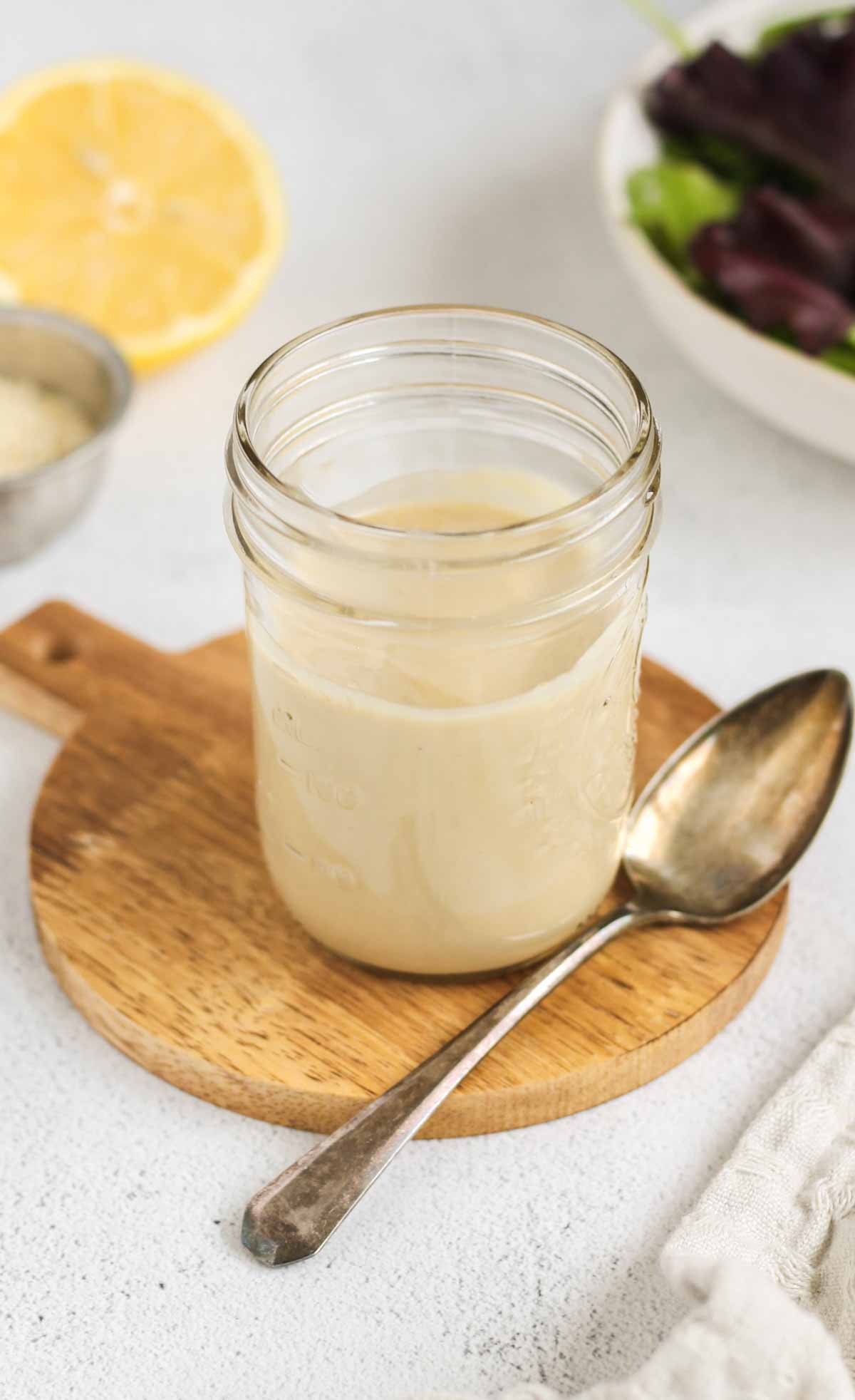 Maple tahini dressing in a mason jar with a spoon on the side of the jar and a lemon and bowl of mixed greens in the background.
