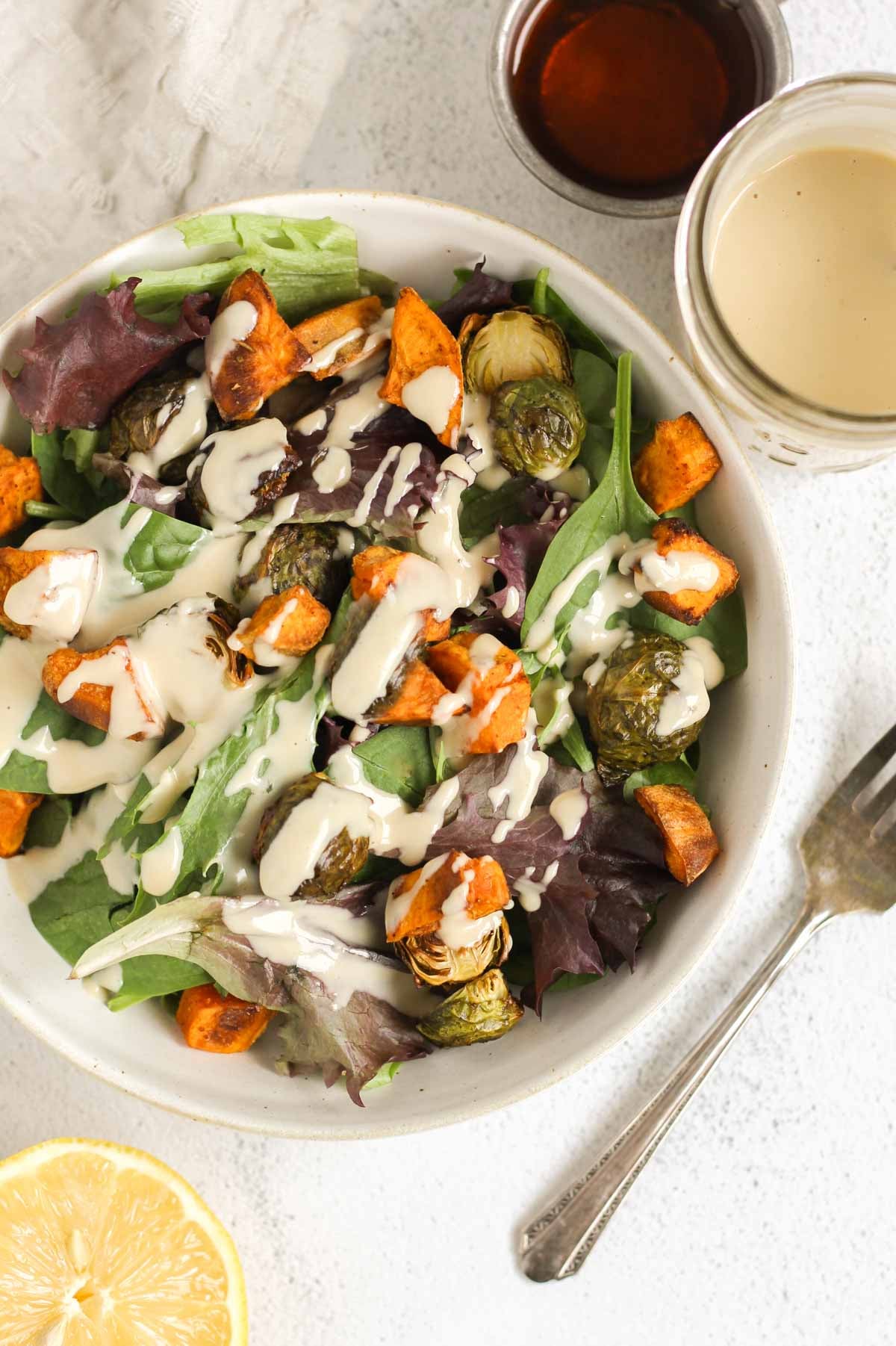 A bowl of greens and roasted veggies with maple tahini dressing drizzled over the top with a fork on the side of the bowl.