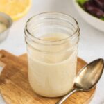 Maple tahini dressing in a mason jar with a spoon on the side of the jar and a lemon and bowl of mixed greens in the background.