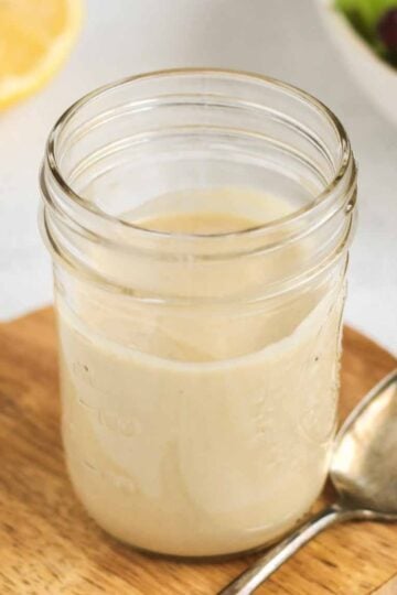 Maple tahini dressing in a mason jar with a spoon on the side of the jar and a lemon and bowl of mixed greens in the background.